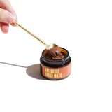 A hand scoops the Urb. Apothecary Artichoke Cacao Mask from its amber glass jar with a golden spoon.