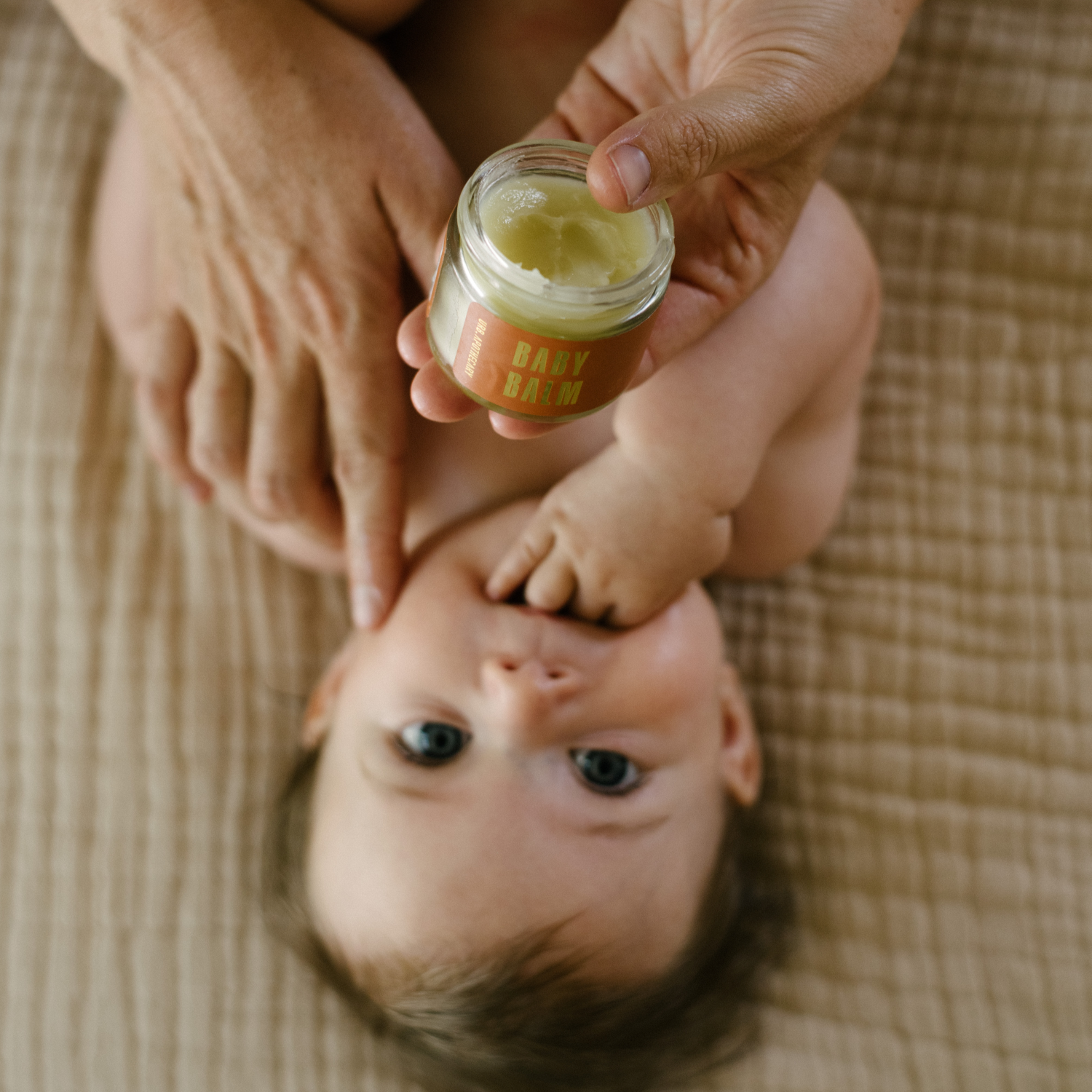 Parent applies organic baby balm to a baby's cheek on a soft blanket.
