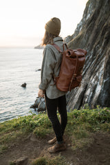 A woman wearing a beanie and leather backpack gazes at the ocean from a grassy cliff during a scenic outdoor adventure.

