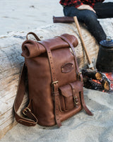 A brown leather roll-top backpack leans against driftwood during a camping trip with a bonfire and coffee brewing nearby.
