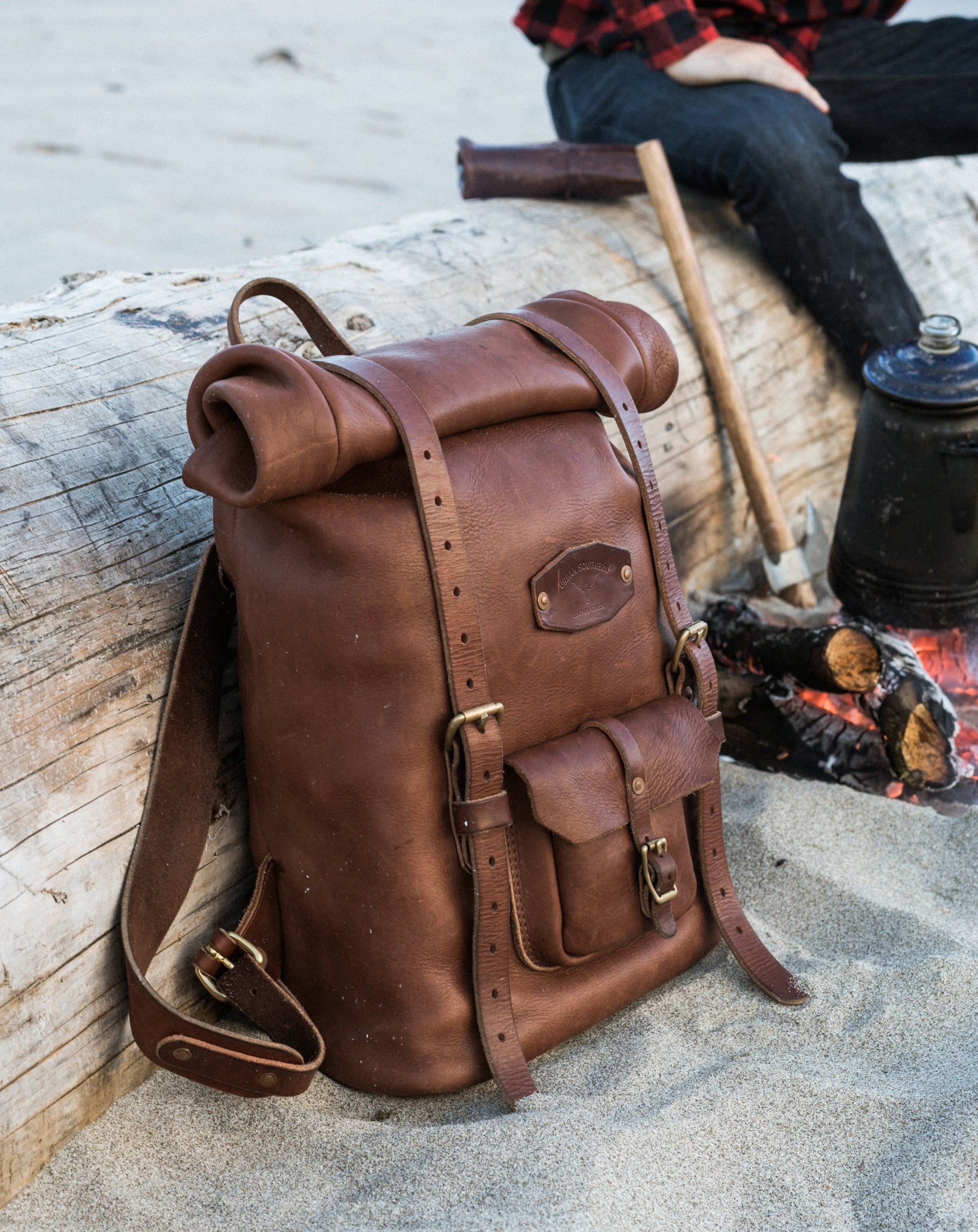 A brown leather roll-top backpack leans against driftwood during a camping trip with a bonfire and coffee brewing nearby.
