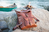 A handcrafted leather backpack sits on a rocky shore next to a canoe and crackling campfire.
