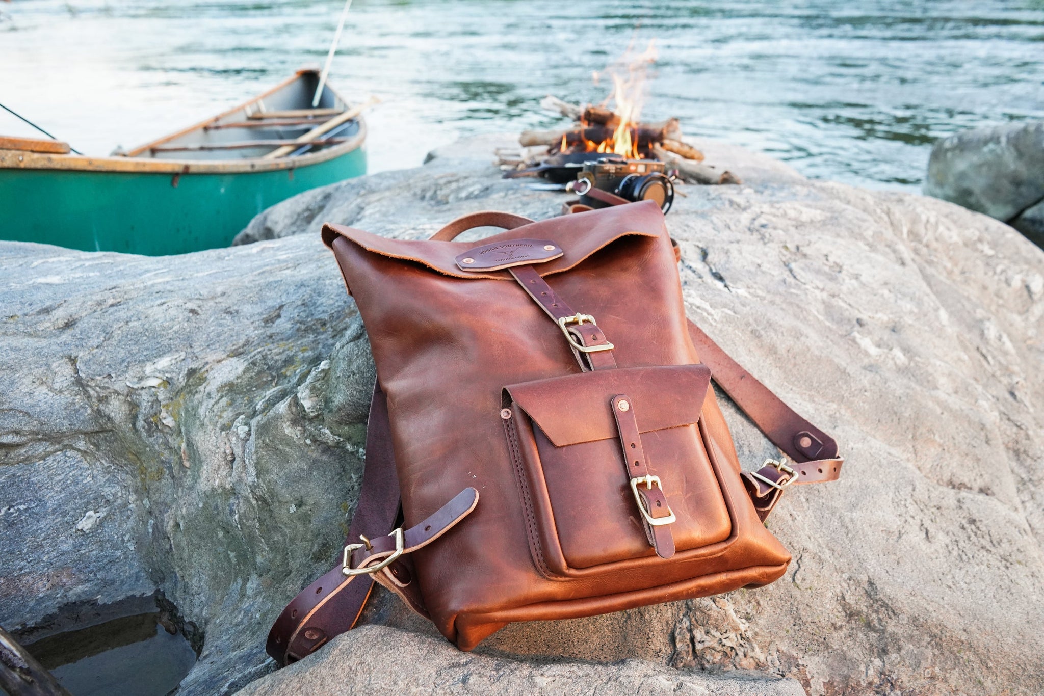 A handcrafted leather backpack sits on a rocky shore next to a canoe and crackling campfire.
