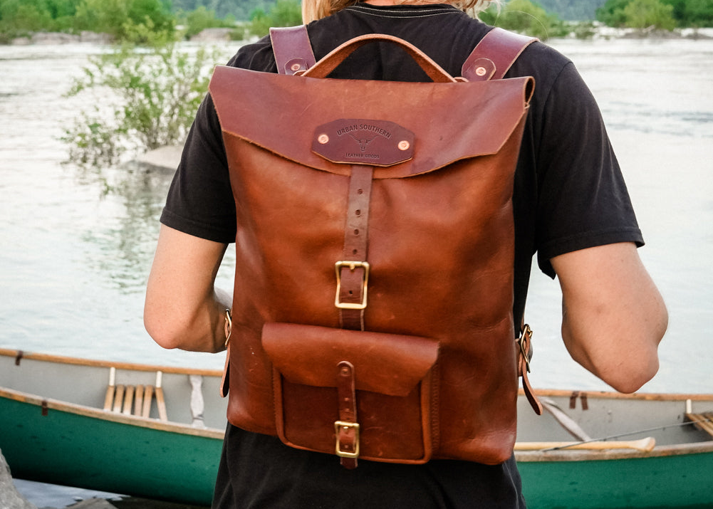 Person with a brown leather backpack standing by a body of water with a canoe.
