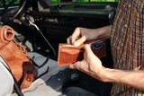 Inside a classic campervan, a man in a yellow plaid shirt opens a brown leather wallet, revealing credit cards.
