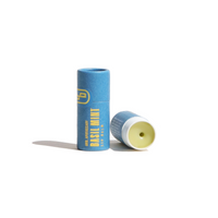Two tubes of URB Apothecary Basil Mint Lip Balm sit on a white surface, one open to show the balm.
