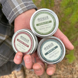 Man holds three tins of Rogue Evergreens beard balm, featuring Douglas-fir, Grand fir, and Incense Cedar scents.
