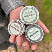 A man shows off three tins of Rogue Evergreens beard balm, including Douglas Fir, Incense Cedar, and Grand Fir varieties.

