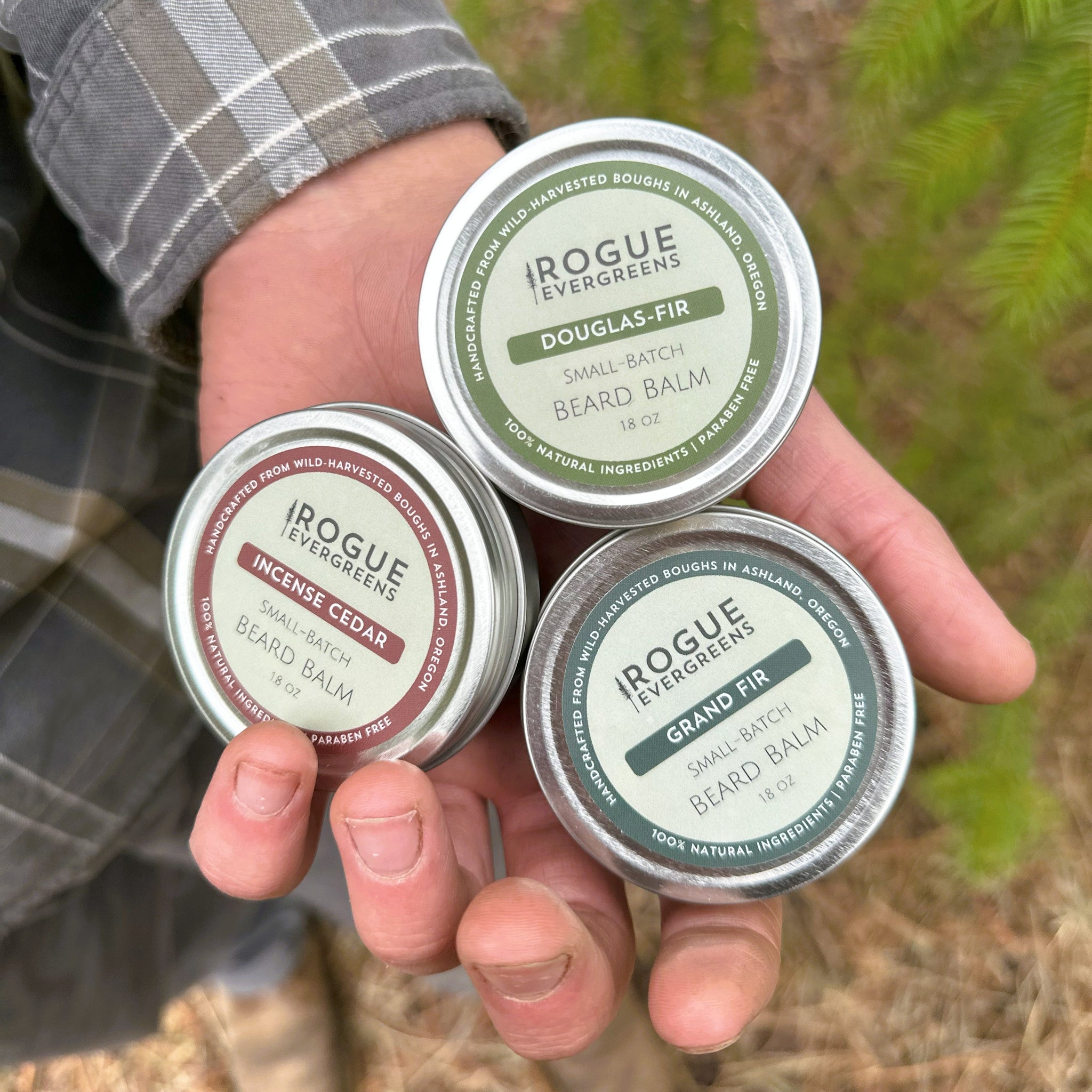 A man shows off three tins of Rogue Evergreens beard balm, including Douglas Fir, Incense Cedar, and Grand Fir varieties.
