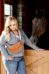 A blonde woman wearing a plaid blazer and brown leather crossbody bag pets her brown horse in its stable.
