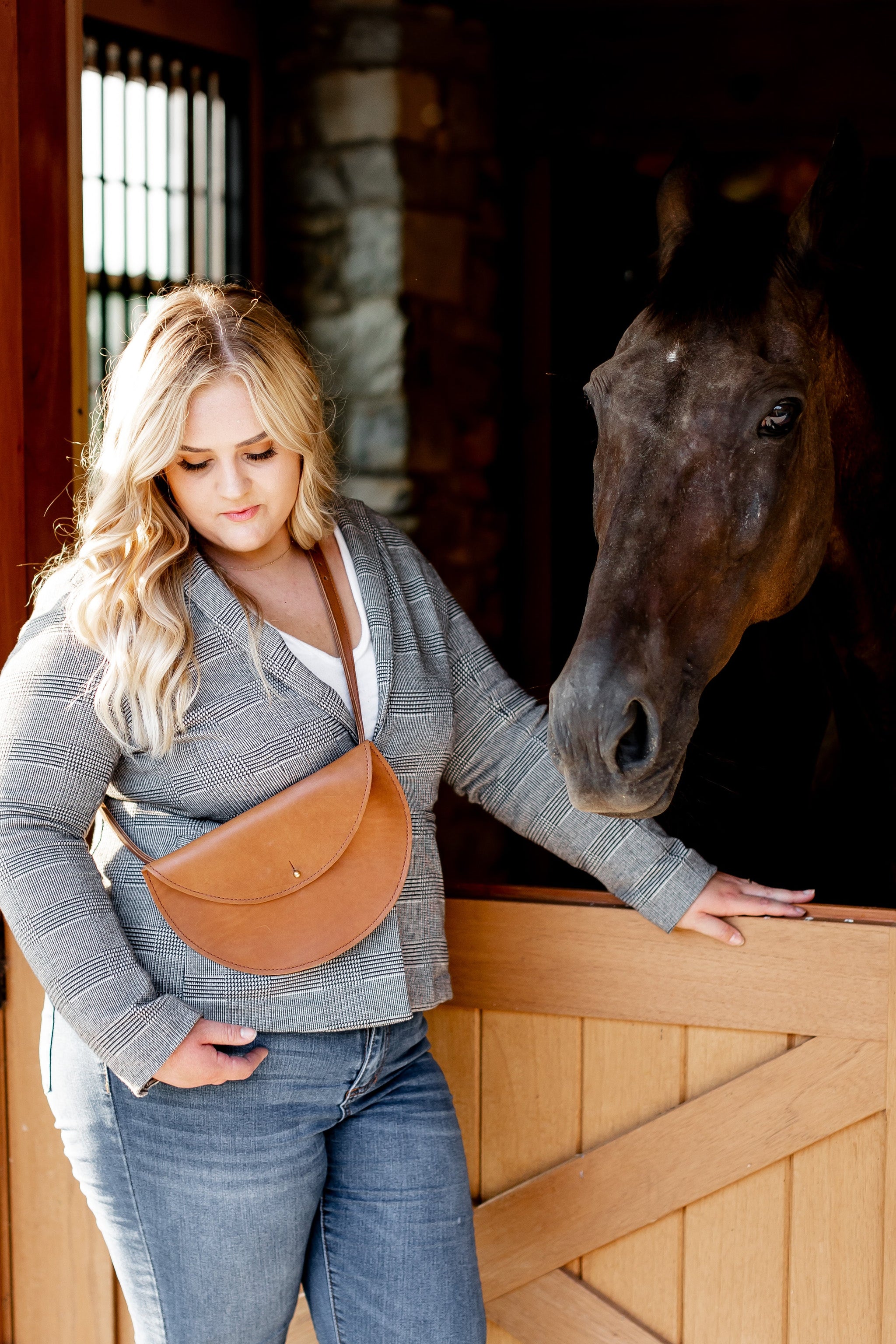 A blonde woman wearing a plaid blazer and brown leather crossbody bag pets her brown horse in its stable.
