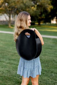 A woman wearing a blue floral dress carries a large, black leather tote bag over her shoulder outdoors.
