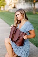 A woman wearing a blue dress looks inside a brown leather tote bag outdoors.