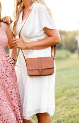 Woman models a stylish brown leather crossbody bag with a white summer dress outdoors.
