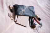 A black leather makeup bag spills out beauty products including brushes, eyeshadow palette, and lip gloss onto a soft pink blanket.
