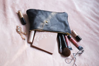 A black leather makeup bag spills out beauty products including brushes, eyeshadow palette, and lip gloss onto a soft pink blanket.
