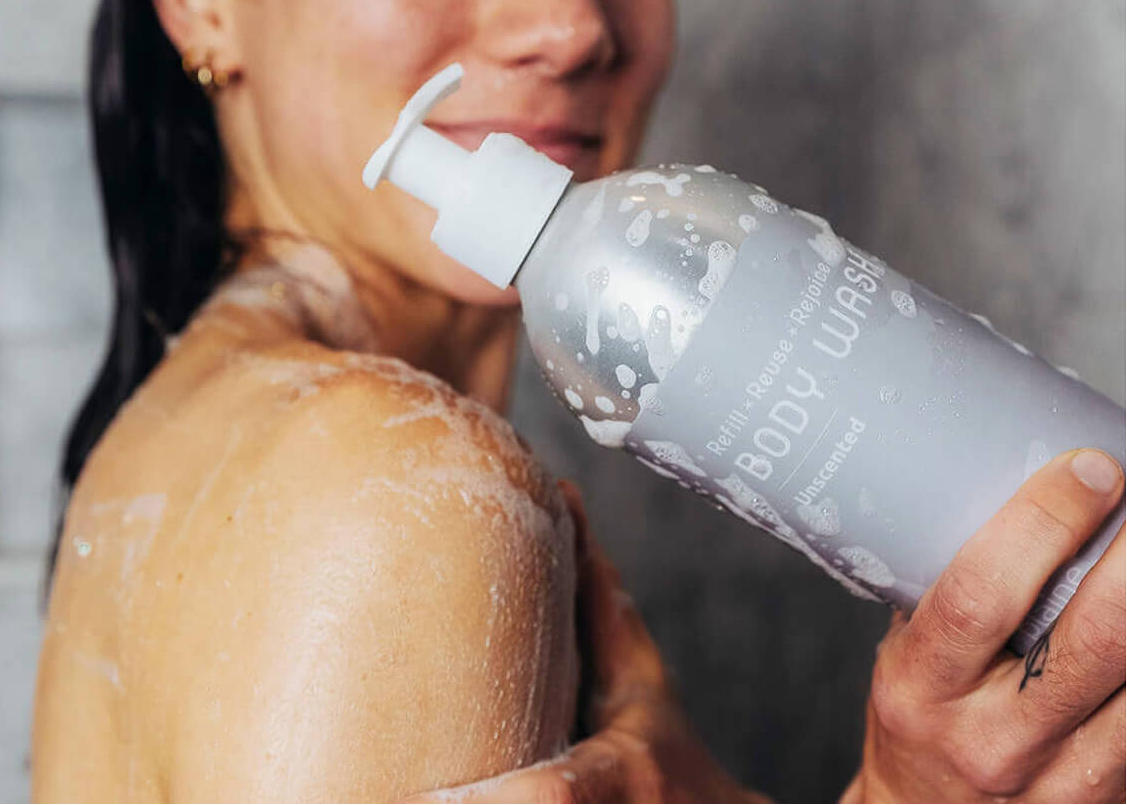 A close-up lifestyle shot of a woman holding a silver body wash bottle with a white pump dispenser, with soap lather visible on her shoulder and the bottle covered in droplets.