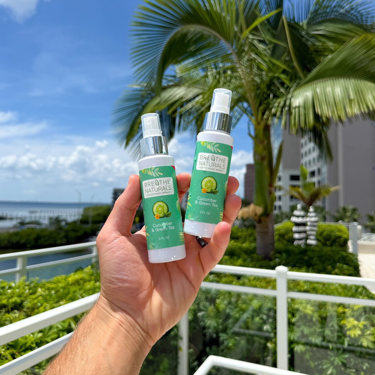 A hand holds two bottles of Breathe Naturals Cucumber & Green Tea deodorant spray against a tropical background.
