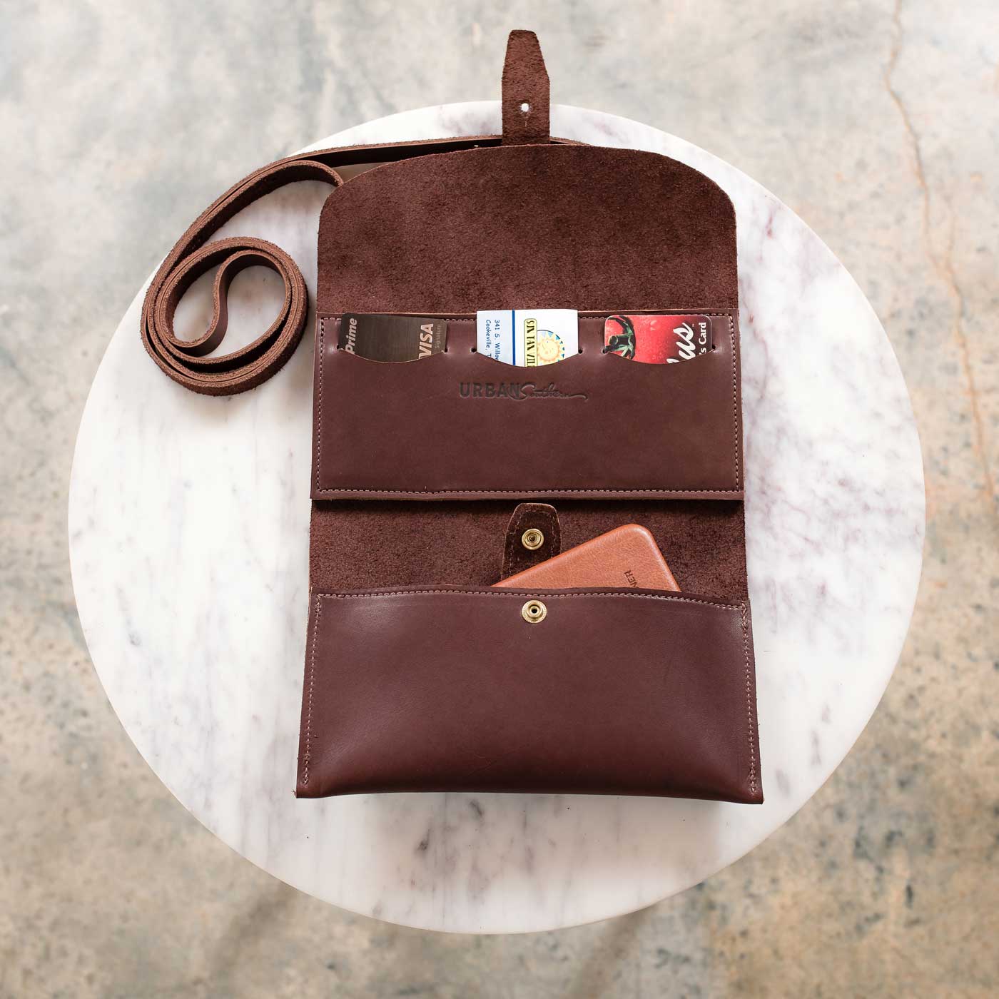 Displaying an open brown leather crossbody wallet reveals credit cards and a phone inside.

