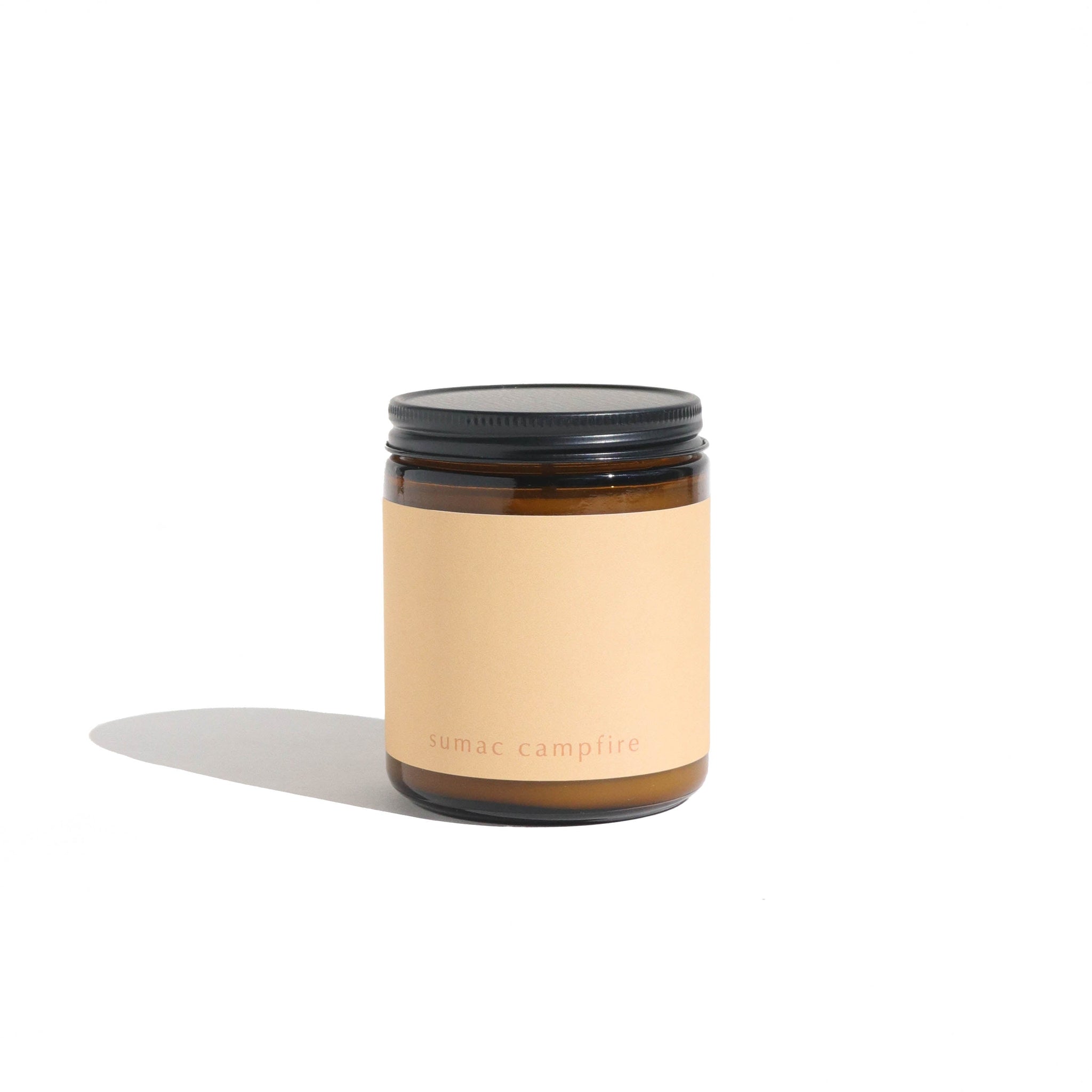 Studio shot presents a Sumac Campfire scented candle, featuring an amber glass jar, minimalist label, and black lid.
