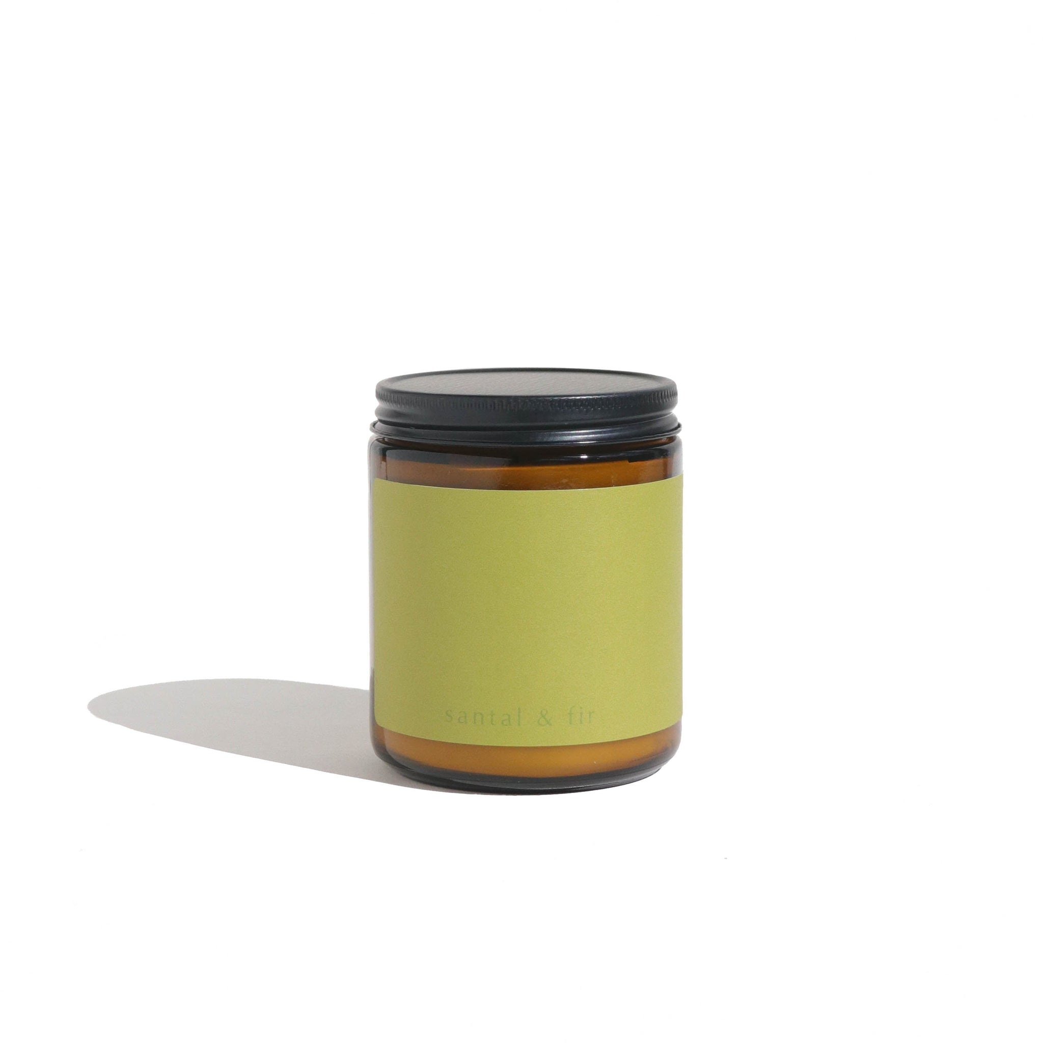 View a santal and fir scented candle in a brown glass jar with a black lid, against a white background.