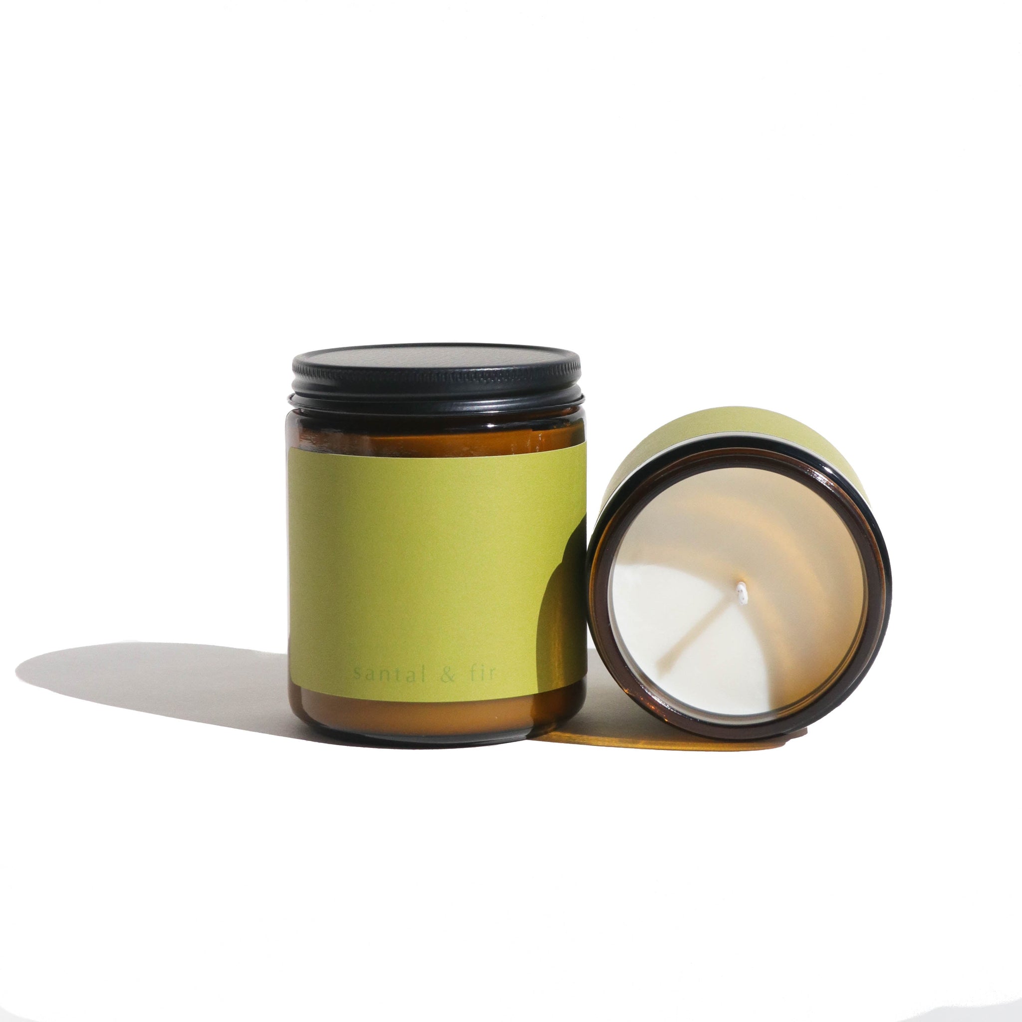Two santal & fir scented candles sit on a white surface, one with its lid on and the other lying on its side showing the wax.
