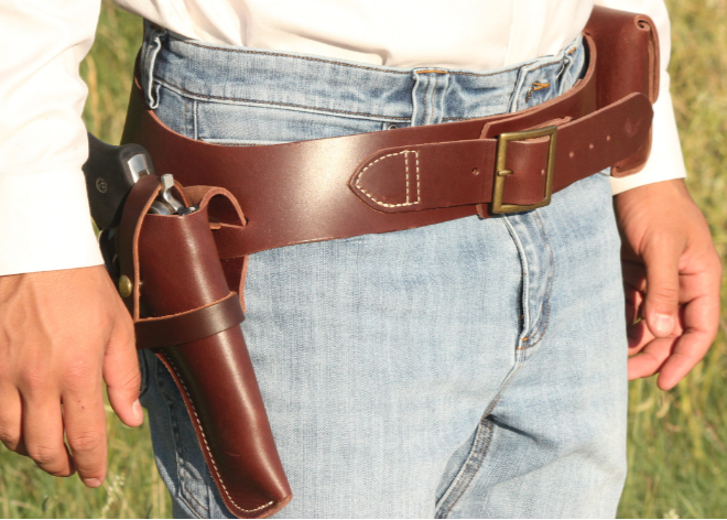 Person wearing a brown leather belt with a holster holding a gun, standing in a grassy field.