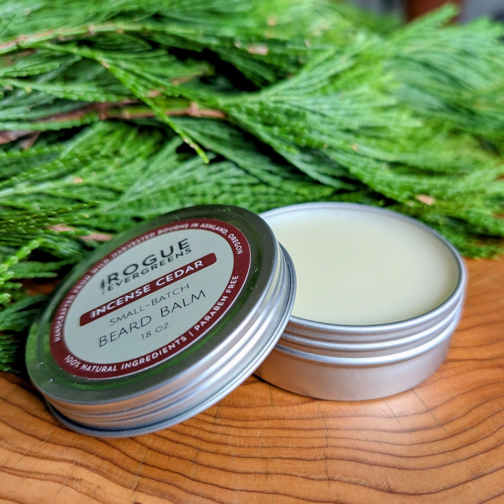 Rogue Evergreens presents their small-batch Incense Cedar beard balm in a silver tin, resting on wooden planks beside fresh evergreen boughs.
