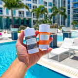 A hand holds two Breathe Naturals deodorant sticks, Cedar Spice and Orange Creamsicle, in front of a sunny resort swimming pool.
