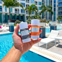A hand holds two Breathe Naturals deodorant sticks, Cedar Spice and Orange Creamsicle, in front of a sunny resort swimming pool.
