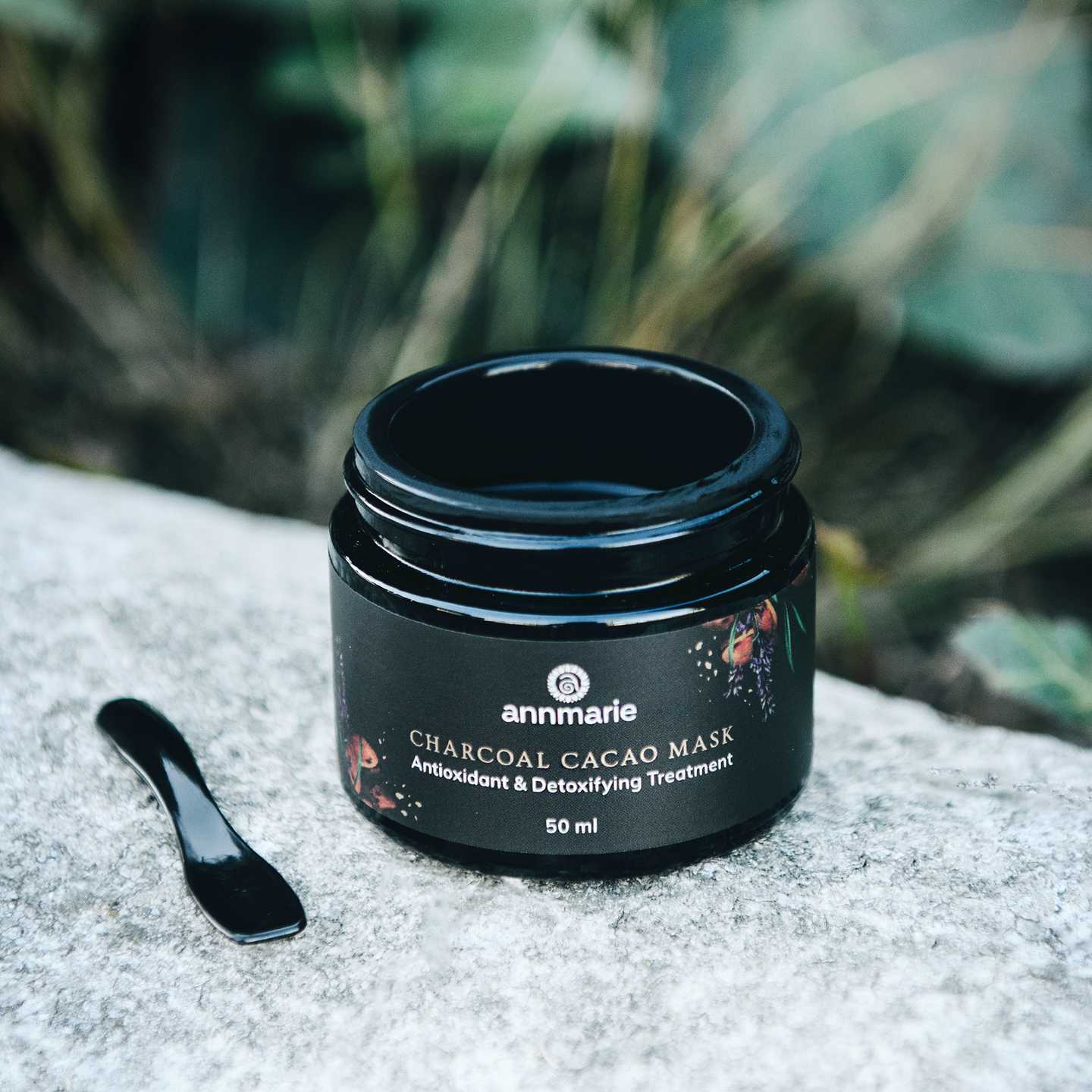 Annmarie's Charcoal Cacao Mask, an antioxidant and detoxifying treatment, sits beside its applicator on a textured gray surface against a blurred green background.
