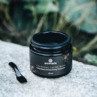 Annmarie's Charcoal Cacao Mask, an antioxidant and detoxifying treatment, sits beside its applicator on a textured gray surface against a blurred green background.
