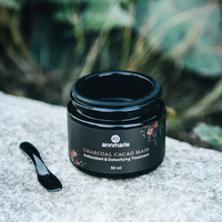 Annmarie's Charcoal Cacao Mask, an antioxidant and detoxifying treatment, sits beside its applicator on a textured gray surface against a blurred green background.

