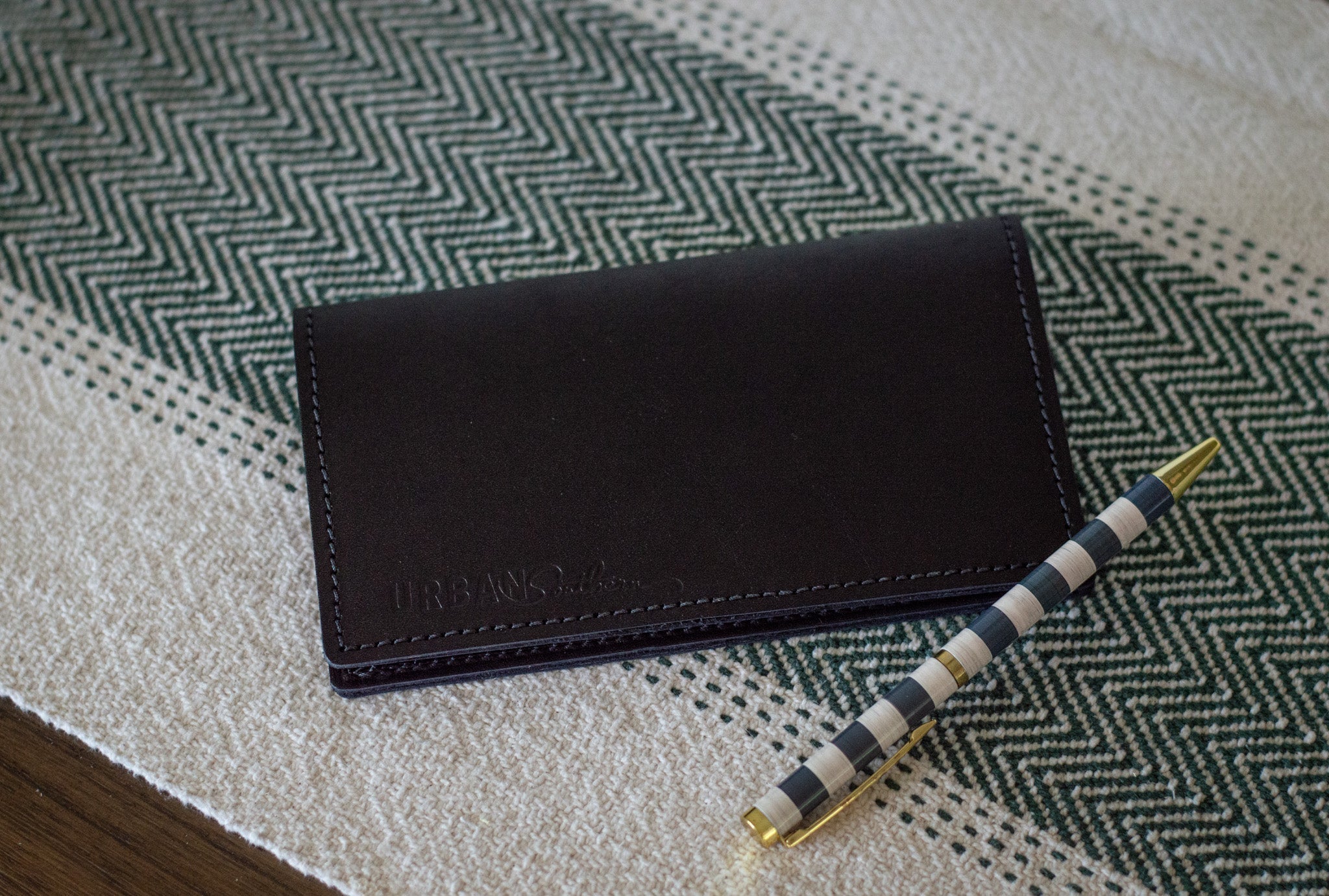 A black leather Urban Status wallet rests beside a stylish blue and white striped pen on a herringbone-patterned surface.
