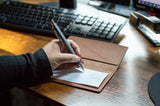 A hand holding a pen writes on a check lying on a brown leather checkbook cover at a wooden desk with office supplies.
