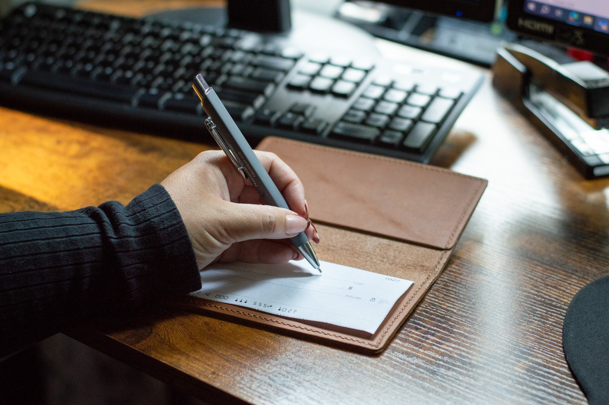 A hand holding a pen writes on a check lying on a brown leather checkbook cover at a wooden desk with office supplies.
