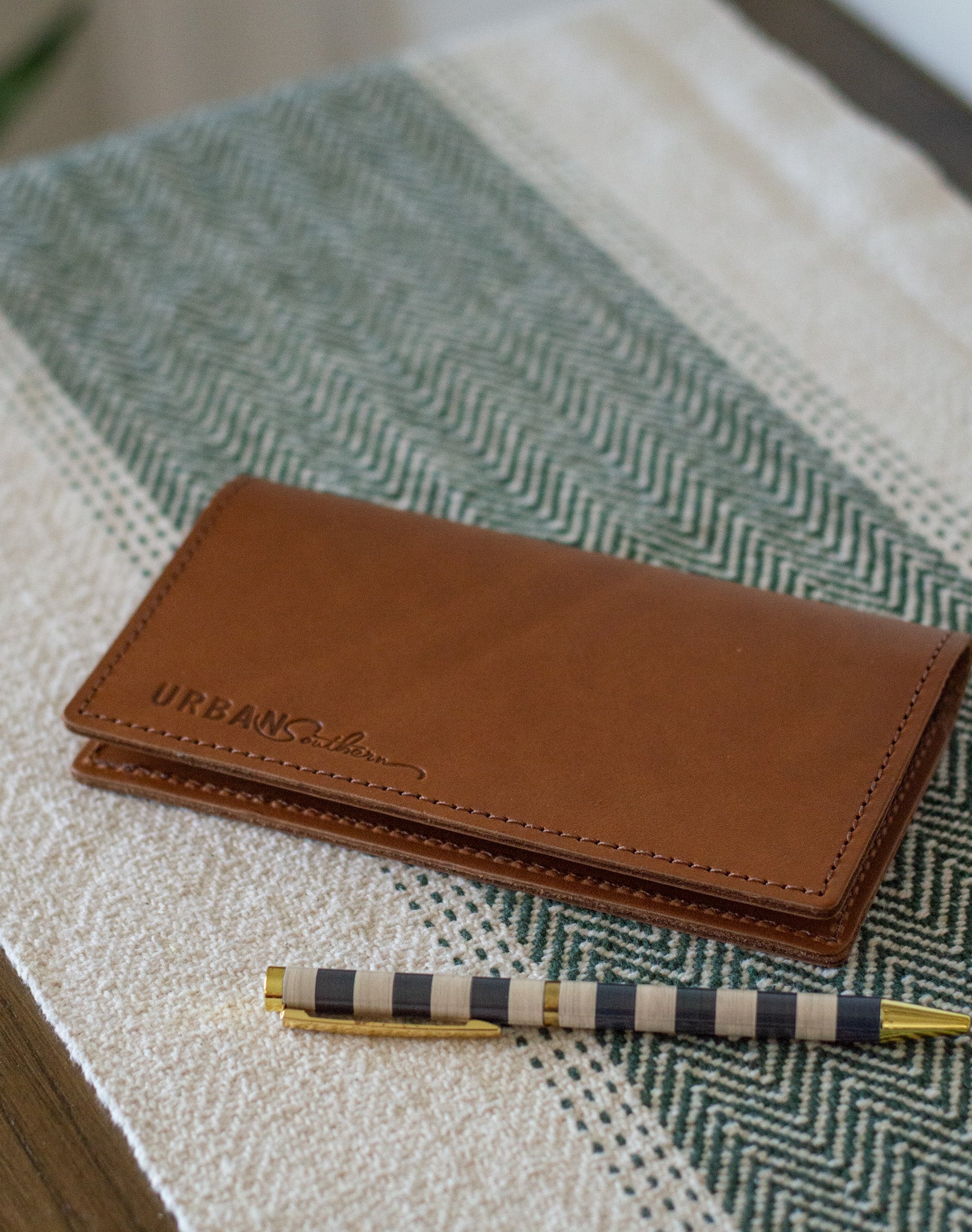 A brown leather wallet with "URBAN Southern" branding rests beside a striped pen on a textured cream and green surface.
