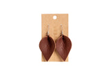 The image shows handcrafted brown leather leaf earrings hanging on a branded cardboard display card.
