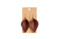 The image shows handcrafted brown leather leaf earrings hanging on a branded cardboard display card.
