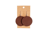 Two brown, circular leather earrings dangle from silver hooks on a branded display card.
