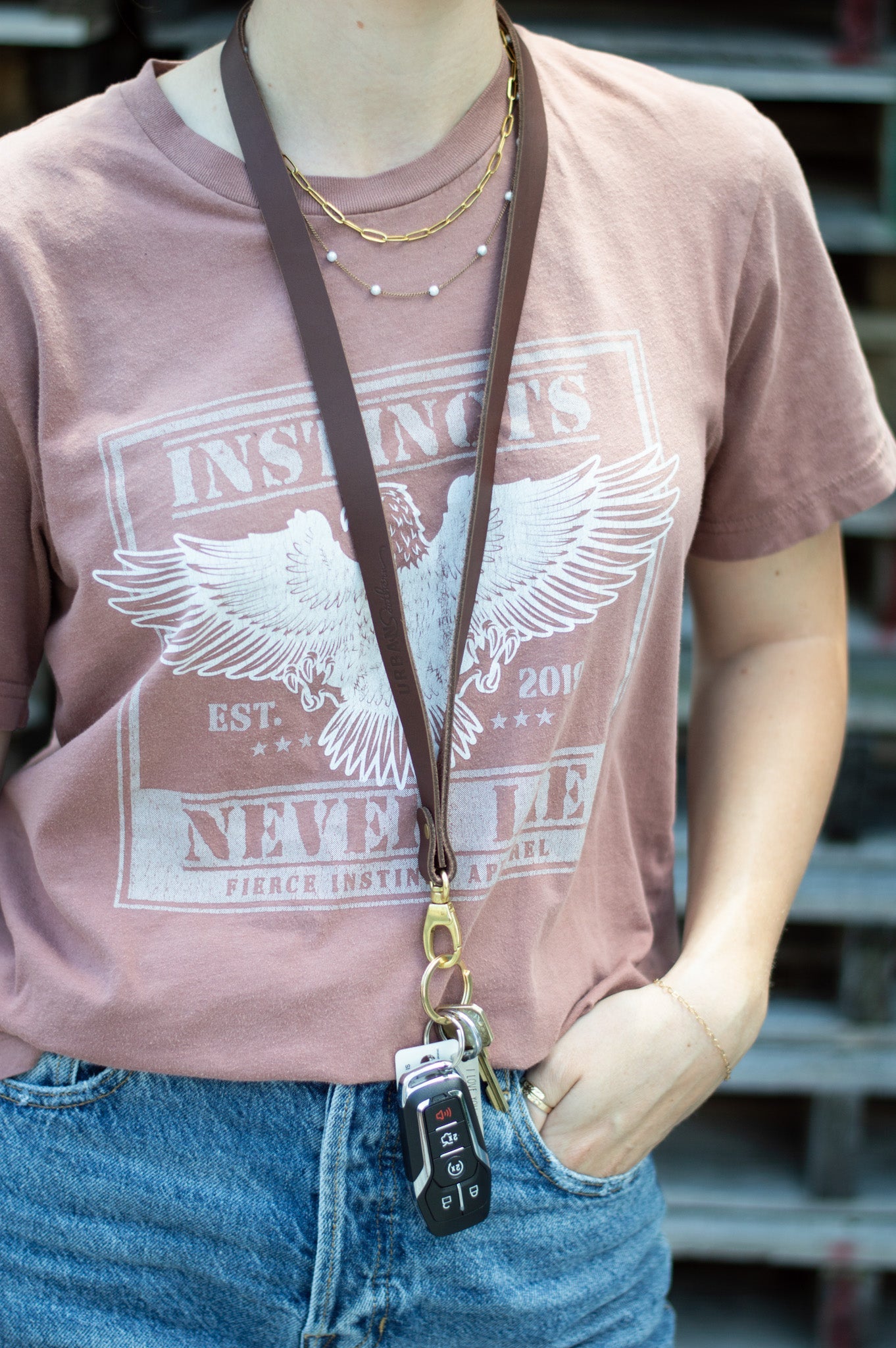 A woman models a stylish brown leather lanyard with keys, a printed mauve t-shirt, layered gold necklaces, and blue jeans.
