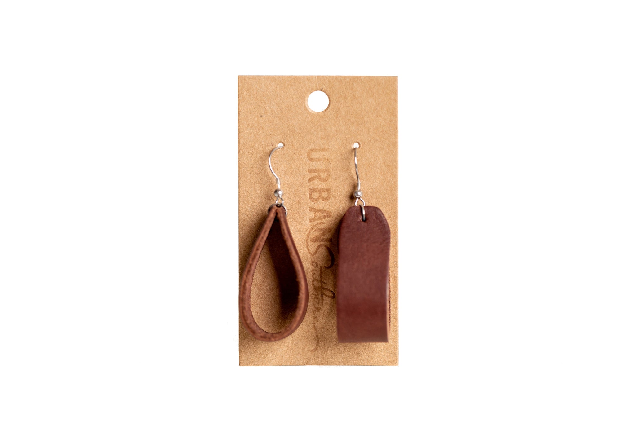 Displays Urban Southern leather earrings, one teardrop and one rectangular, hanging on a branded kraft paper card.
