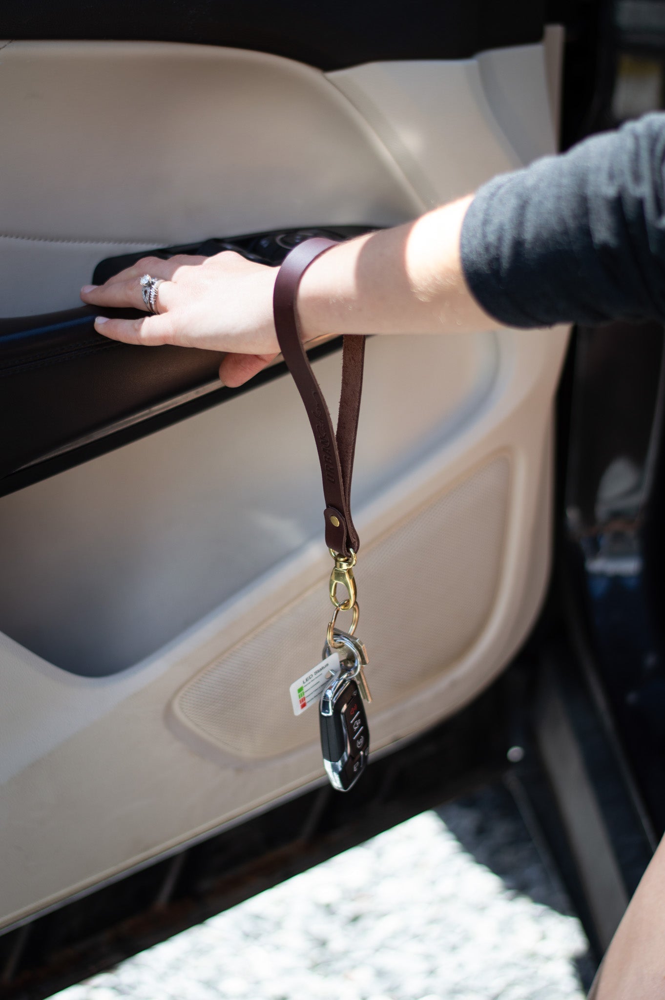 A woman's hand, adorned with a diamond ring, holds a brown leather key lanyard featuring a car key while inside a vehicle with a light-colored interior.
