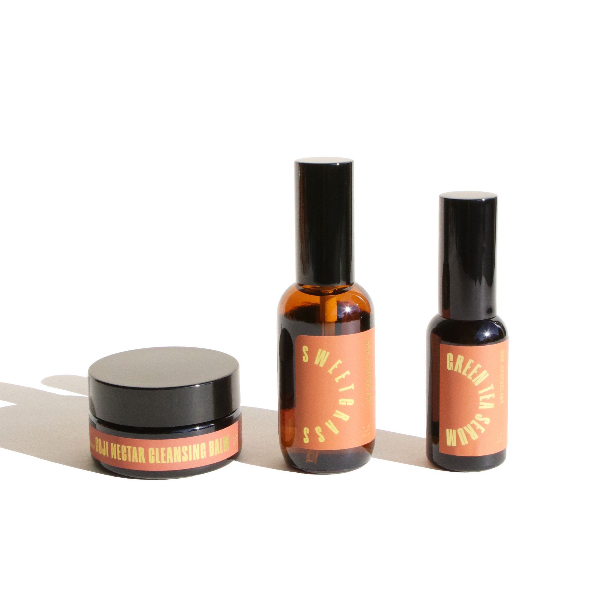 Showcases Urb Apothecary's Goji Nectar Cleansing Balm, Sweetgrass Oil, and Green Tea Serum on a white surface.
