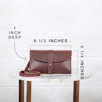 The image showcases the dimensions of a brown leather crossbody bag, displaying its 8 1/2 inch width, 5 1/4 inch height, and 1 inch depth on a marble-topped table.

