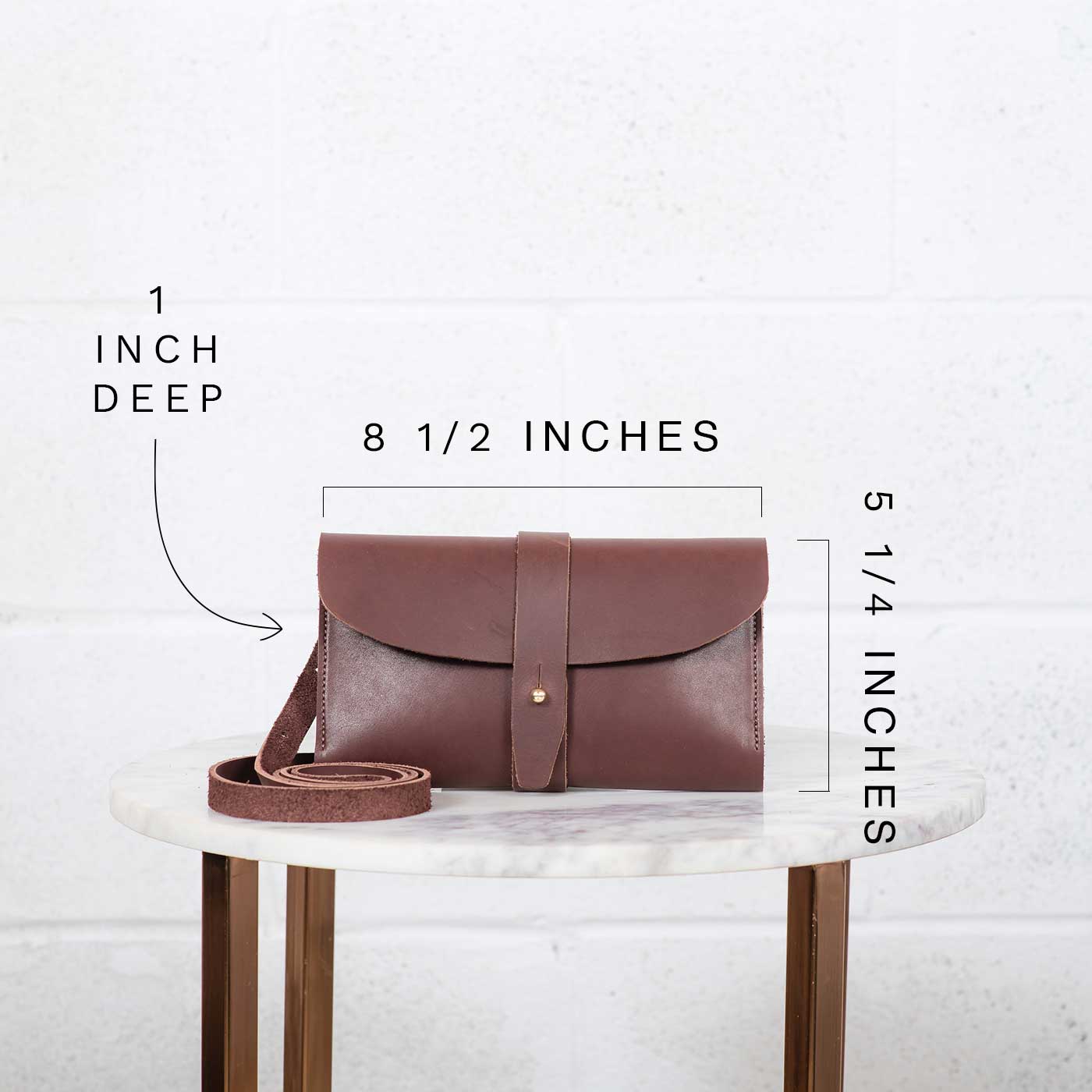 The image showcases the dimensions of a brown leather crossbody bag, displaying its 8 1/2 inch width, 5 1/4 inch height, and 1 inch depth on a marble-topped table.
