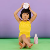 A young girl in a yellow swimsuit sits cross-legged, balancing a white object on her head near a bottle of Dabble & Dollop Coconut Gel kids' bath product.
