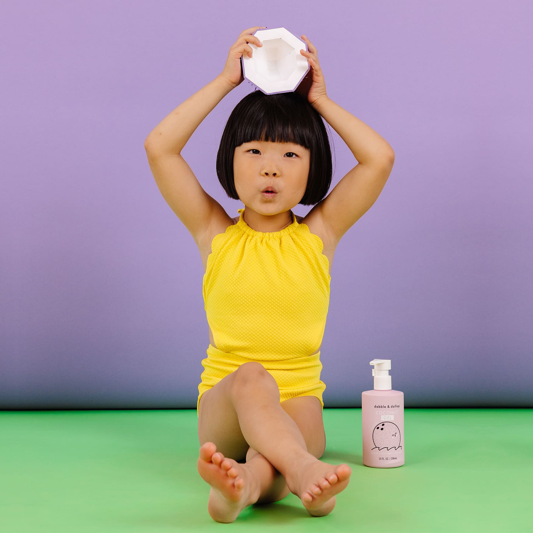 A young girl in a yellow swimsuit sits cross-legged, balancing a white object on her head near a bottle of Dabble & Dollop Coconut Gel kids' bath product.
