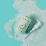 Dabble & Dollop Honeydew Gel body wash bottle sits in a pool of bubbly foam against a bright turquoise background.
