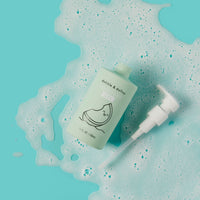 Dabble & Dollop Honeydew Gel body wash bottle sits in a pool of bubbly foam against a bright turquoise background.
